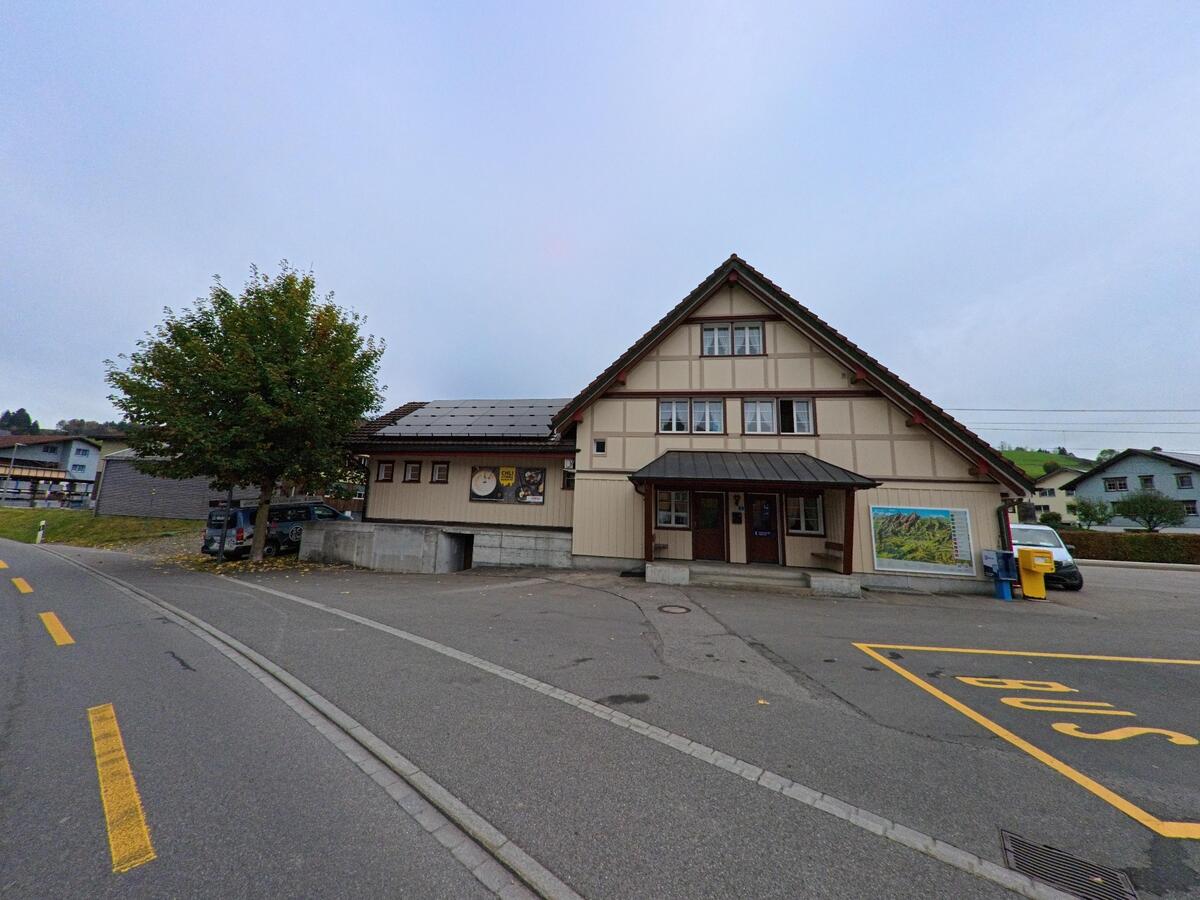 An einer Strasse in Schwende (9057, Kanton Appenzell Innerrhoden) steht ein traditionelles Gebäude mit Fachwerkelementen neben einer Bushaltestelle, Informationstafel und Briefkasten.