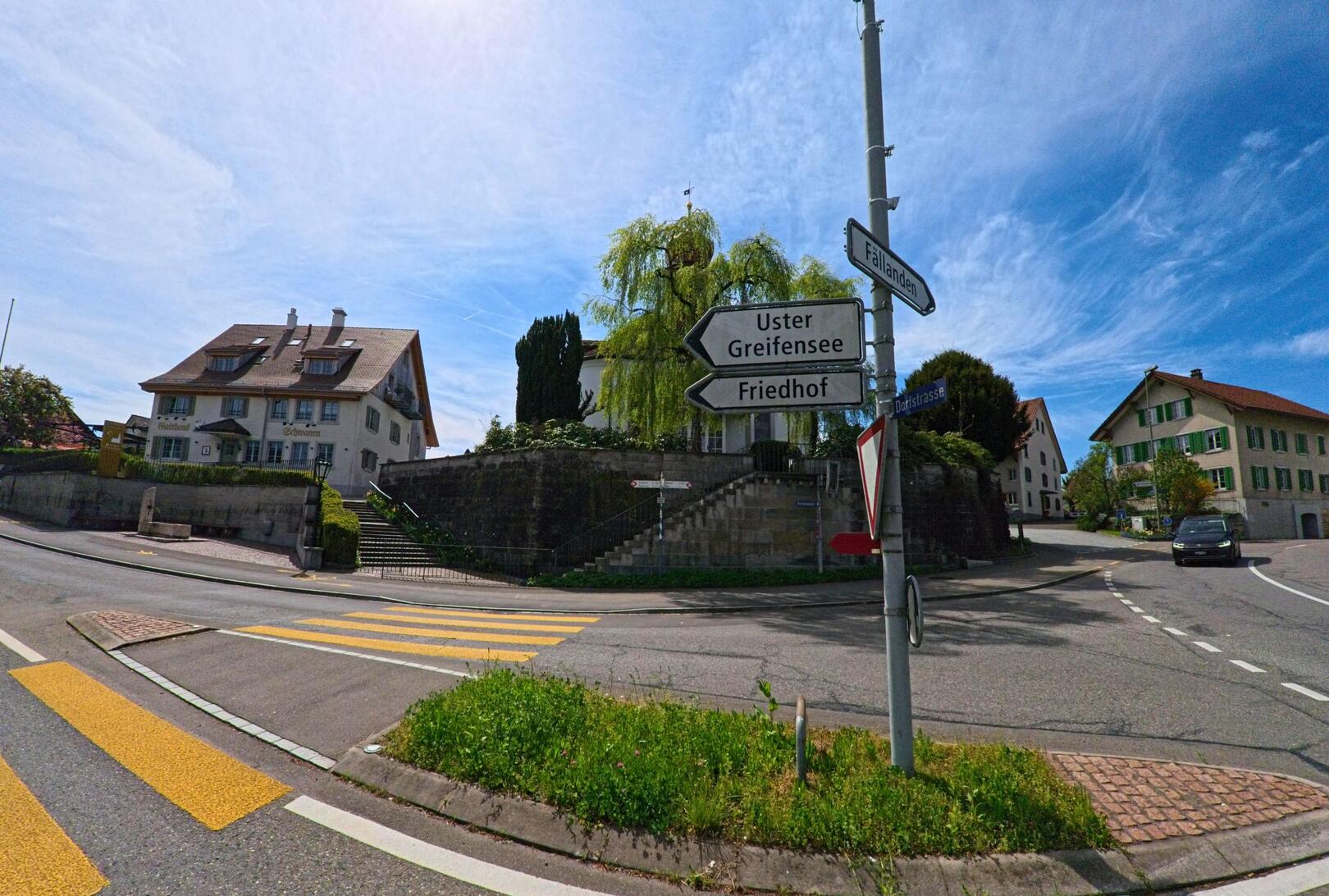An der Dorfstrasse in Schwerzenbach (8603, Kanton Zürich) treffen mehrere Strassen aufeinander, flankiert von traditionellen Wohnhäusern und einer Wegweisung nach Uster, Greifensee und Fällanden.