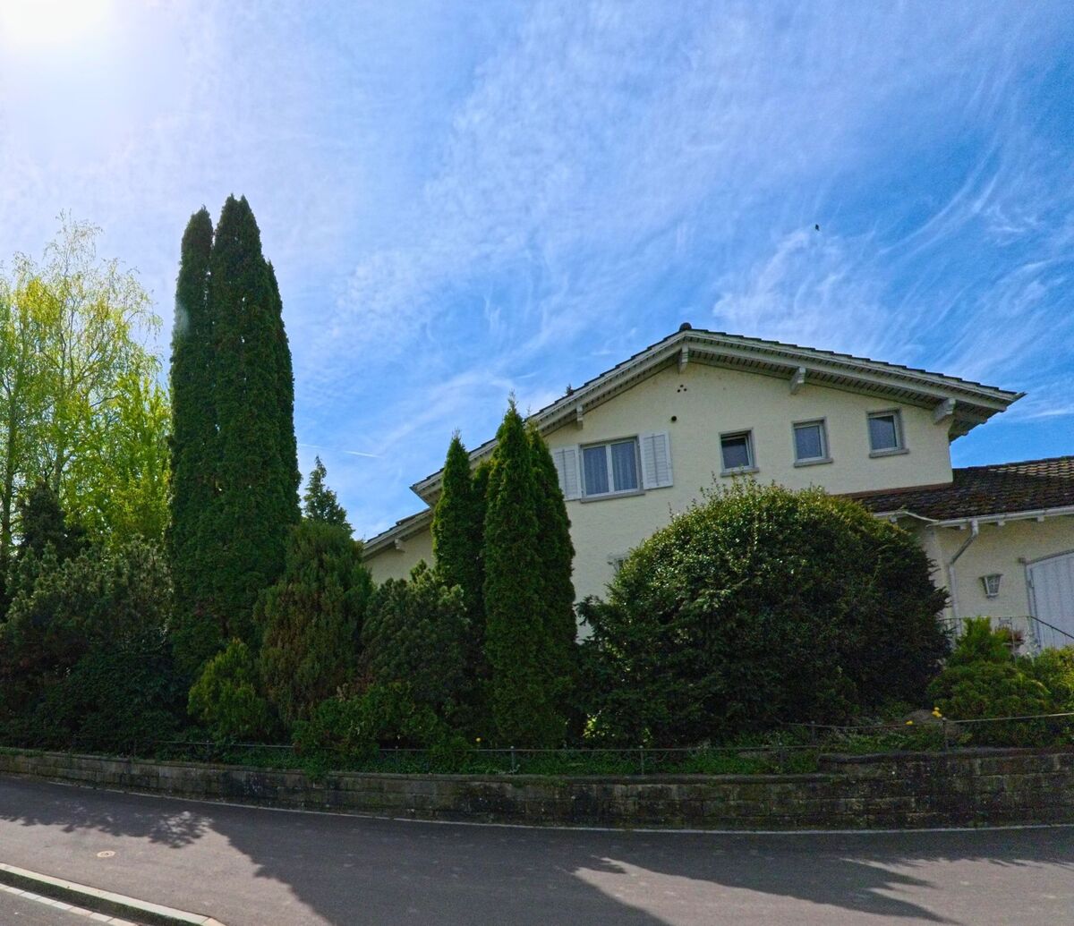 In Schwerzenbach (Kanton Zürich, PLZ 8603) ragt ein helles Wohnhaus mit markantem Dach hinter dicht gewachsenen Zypressen und Sträuchern hervor.