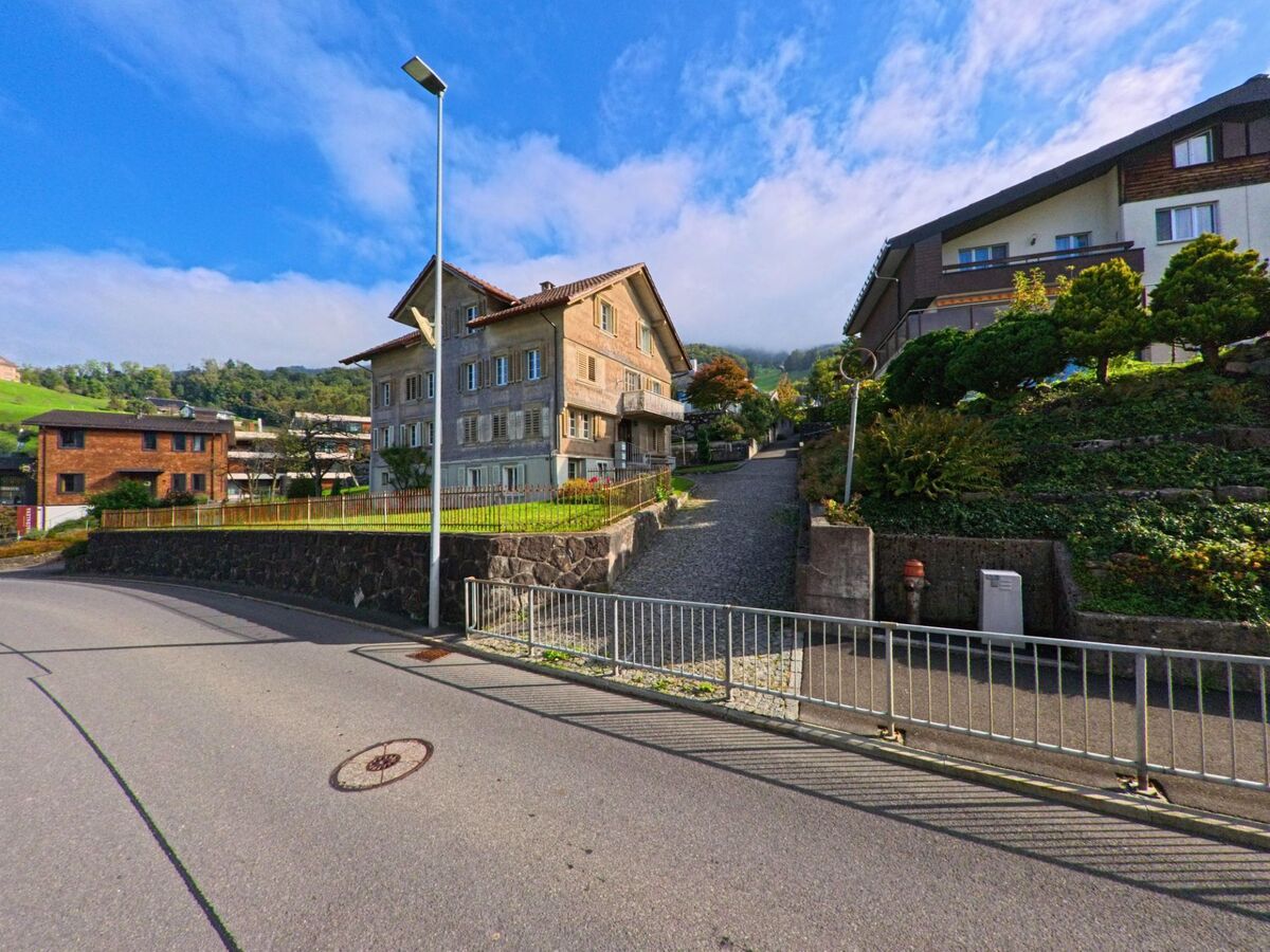 Hinter einem alten Wohnhaus in Schwyz (6423, Kanton Schwyz) fuehrt ein gepflasterter Weg bergauf, umgeben von gepflegten Gaerten und Hanganlagen.