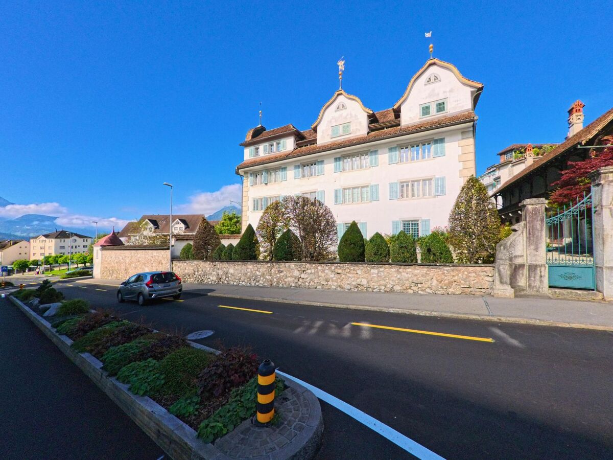 Entlang einer Strasse in Schwyz (6423, Kanton Schwyz) steht ein herrschaftliches Haus mit geschwungenen Giebeln hinter einer Steinmauer und gepflegten Straeuchern.