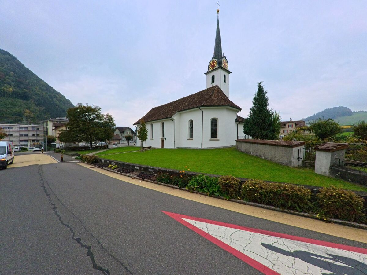 Neben einer kleinen weissen Kirche in Schwyz (6423, Kanton Schwyz) verlaeuft eine ruhige Strasse, eingerahmt von Wiesen und umliegenden Haengern.