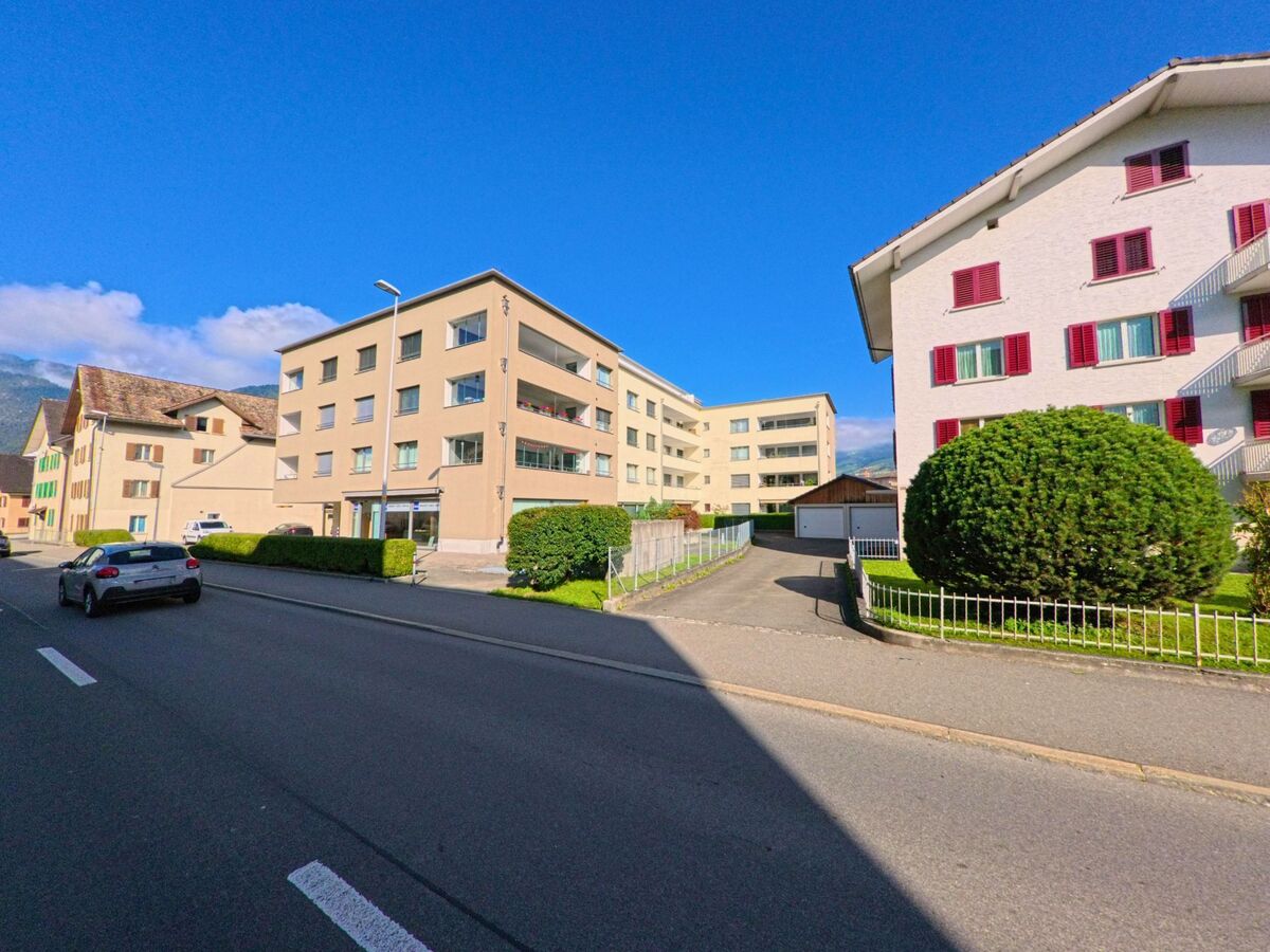 Zwischen modernen Wohngebaeuden in Schwyz (6423, Kanton Schwyz) verlaeuft eine breite Strasse, gesaeumt von gepflegten Hecken und einem kleinen Zufahrtsweg.