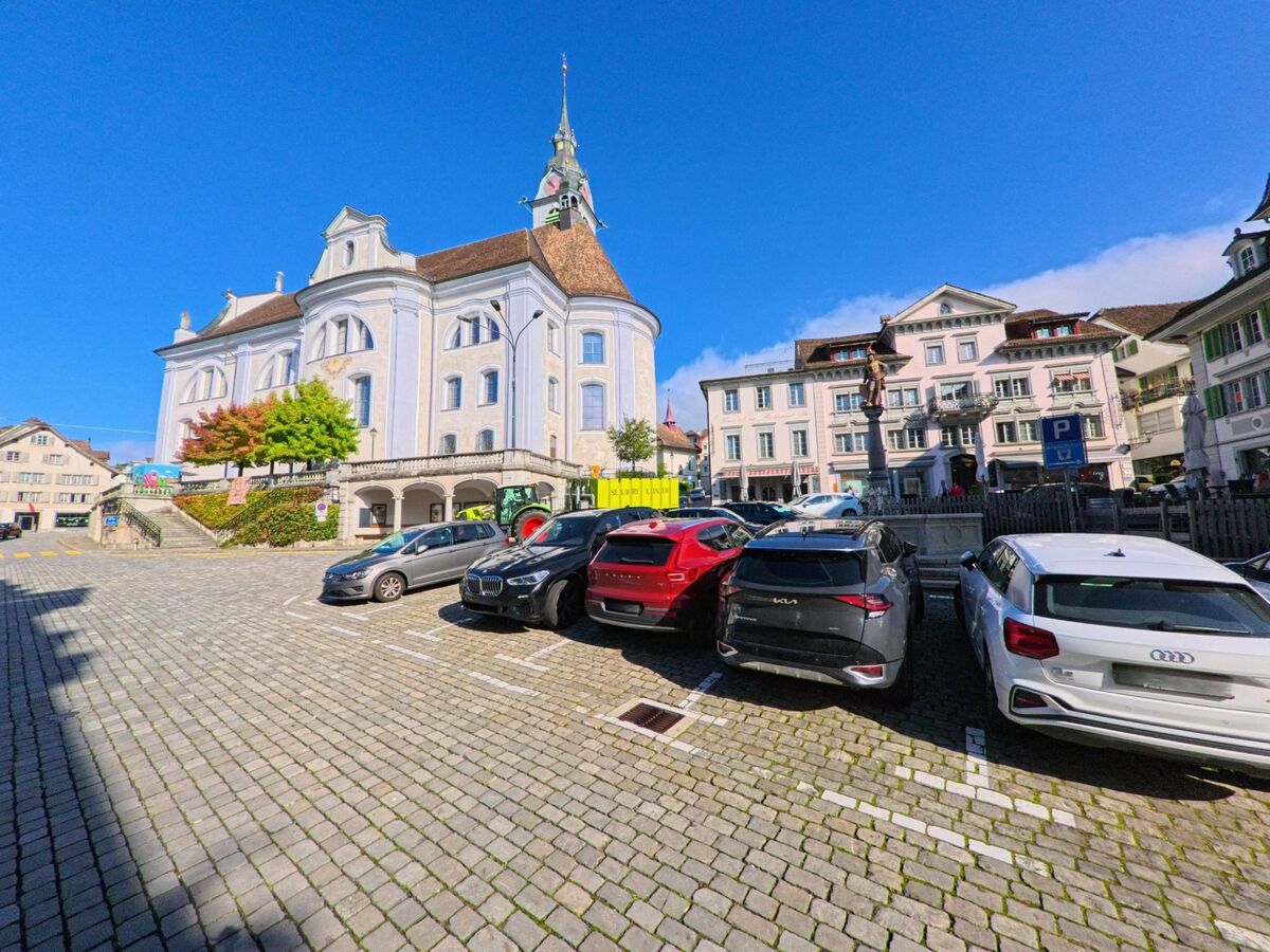 Auf dem gepflasterten Platz in Schwyz (6423, Kanton Schwyz) stehen mehrere Autos vor dem markanten Barockbau und den umliegenden historischen Hausern.