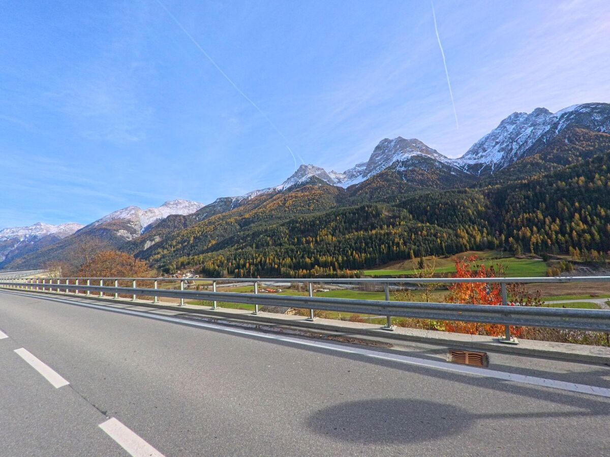 Von einer Strasse mit Leitplanke eröffnet sich in Scuol (7550, Kanton Graubünden) ein weiter Blick über das Tal zu bewaldeten Berghängen und schneebedeckten Gipfeln.