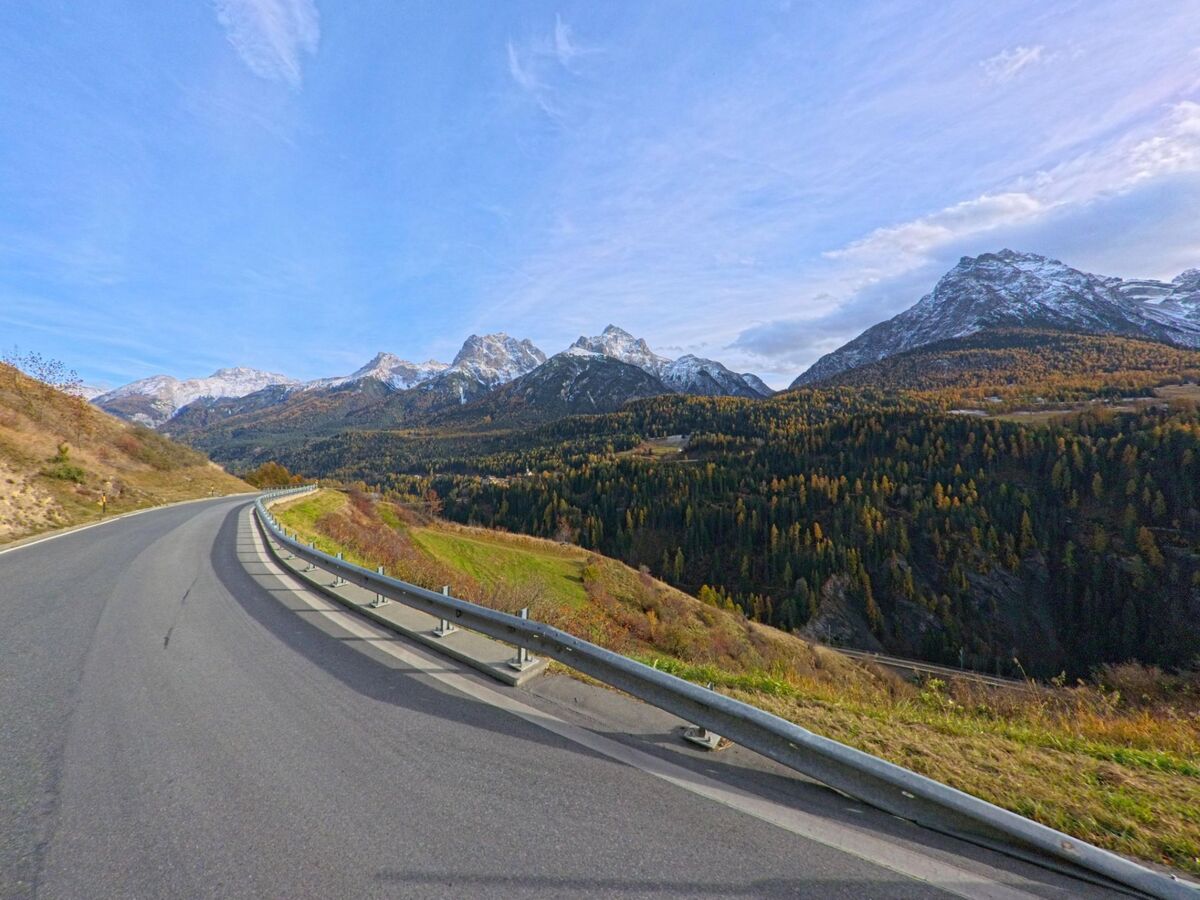 Entlang einer leicht geschwungenen Bergstrasse verläuft in Scuol (7551, Kanton Graubünden) der Blick über bewaldete Hänge hin zu schneebedeckten Gipfeln.