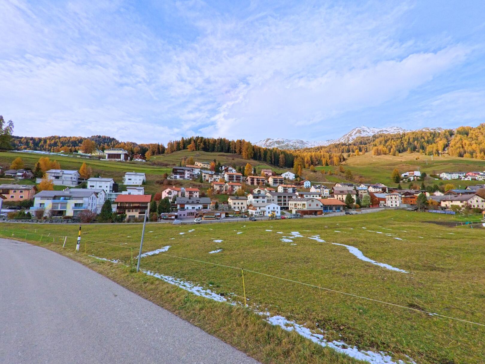 Über offene Wiesen hinweg zeigt sich in Scuol (7551, Kanton Graubünden) ein Dorfteil mit dicht stehenden Wohnhäusern am Hang vor herbstlich gefärbten Wäldern.