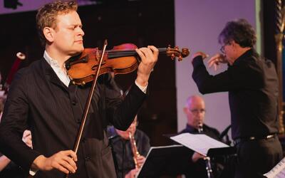 Bei einem Konzert in Fisibach (5467, Kanton Aargau) spielt ein Geiger im Vordergrund, während ein Dirigent das Ensemble leitet.