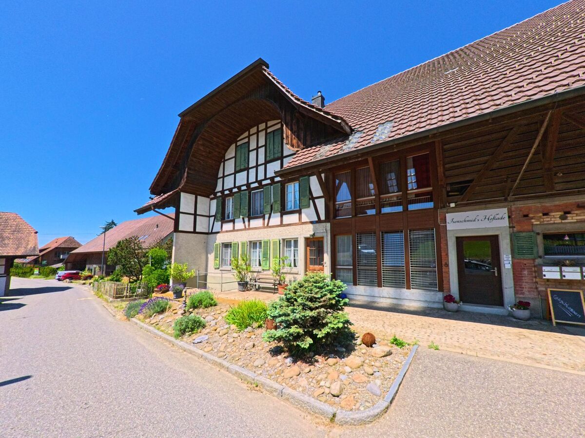In Seedorf (3067, Kanton Bern) steht ein traditionelles Bauernhaus mit grossem Walmdach und grün gestrichenen Fensterläden an einer ruhigen Dorfstrasse, daneben befindet sich ein Hofladen mit Eingang und Schild.