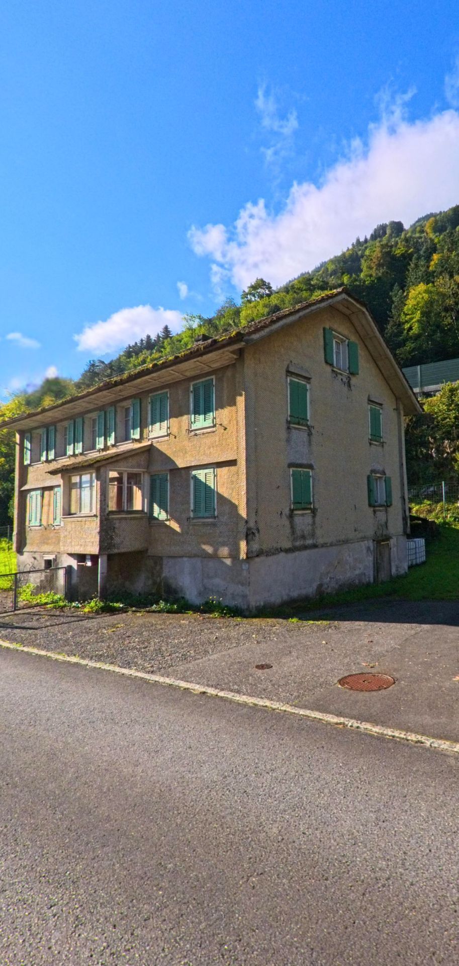 Am Rand einer schmalen Strasse steht ein älteres Wohnhaus mit grünen Fensterläden in Seedorf (6462, Kanton Uri) vor einem bewaldeten Hang.