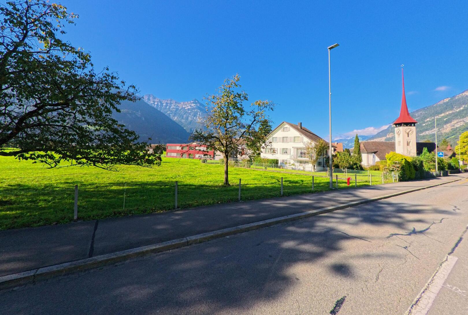 Entlang einer ruhigen Strasse mit Blick auf Wiesen, Wohnhäuser und den markanten Kirchturm zeigt sich das Dorfbild von Seedorf (6462, Kanton Uri) vor alpiner Bergkulisse.