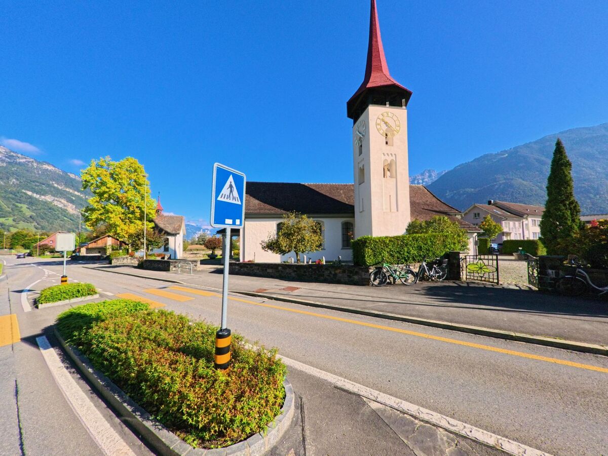 An einem Fussgängerstreifen erhebt sich der markante Kirchturm neben der Strasse im Ortszentrum von Seedorf (6462, Kanton Uri) vor alpiner Kulisse.