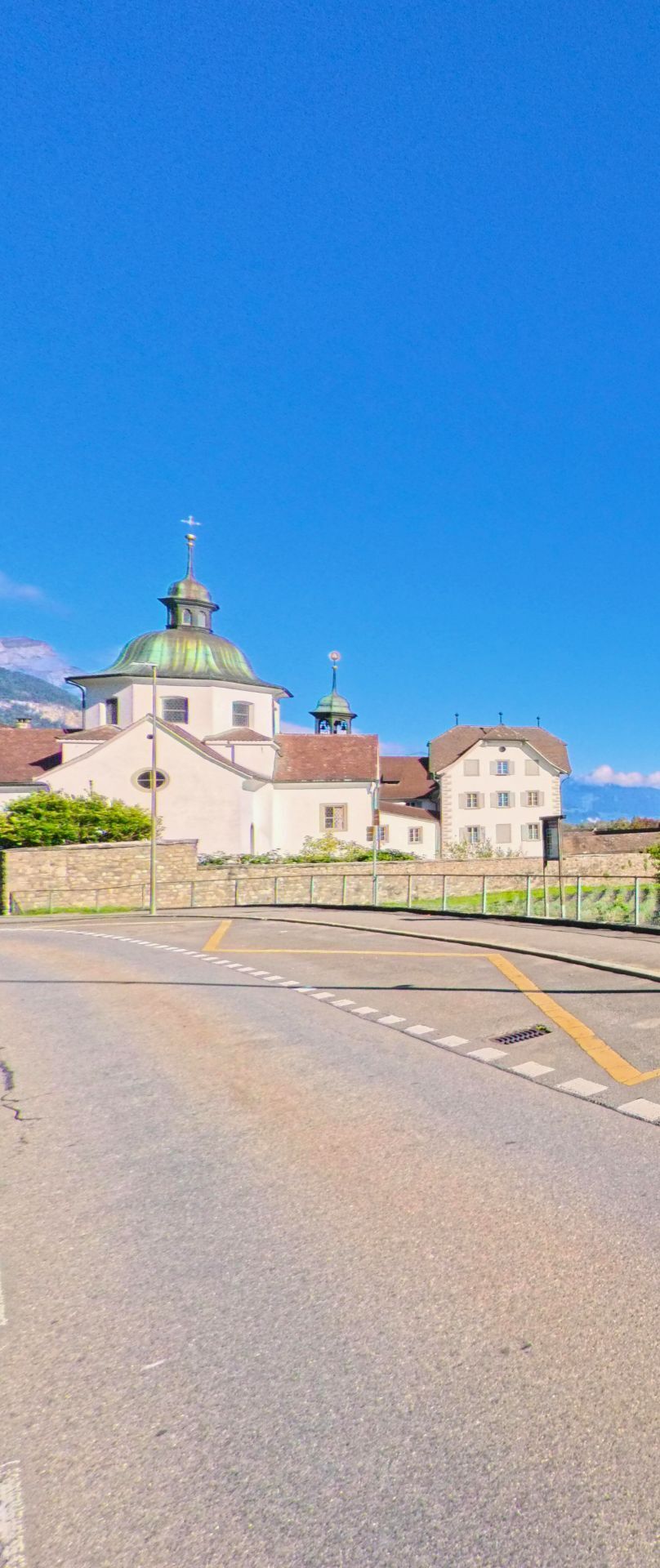 Hinter einer sanft geschwungenen Strasse ist eine historische Kirchenanlage mit markanter Kuppel in Seedorf (6462, Kanton Uri) unter weitem blauem Himmel zu sehen.