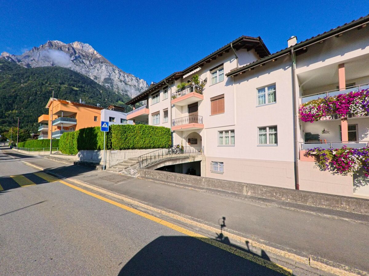 Entlang einer Wohnstrasse mit gepflegten Mehrfamilienhäusern und blühenden Balkonen zeigt sich Seedorf (6462, Kanton Uri) vor einer eindrücklichen Bergkulisse.
