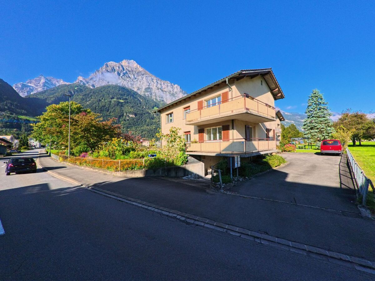 An einer ruhigen Seitenstrasse steht ein Mehrfamilienhaus mit Balkonen in Seedorf (6462, Kanton Uri), dahinter ragen bewaldete Hänge und ein markanter Berggipfel auf.