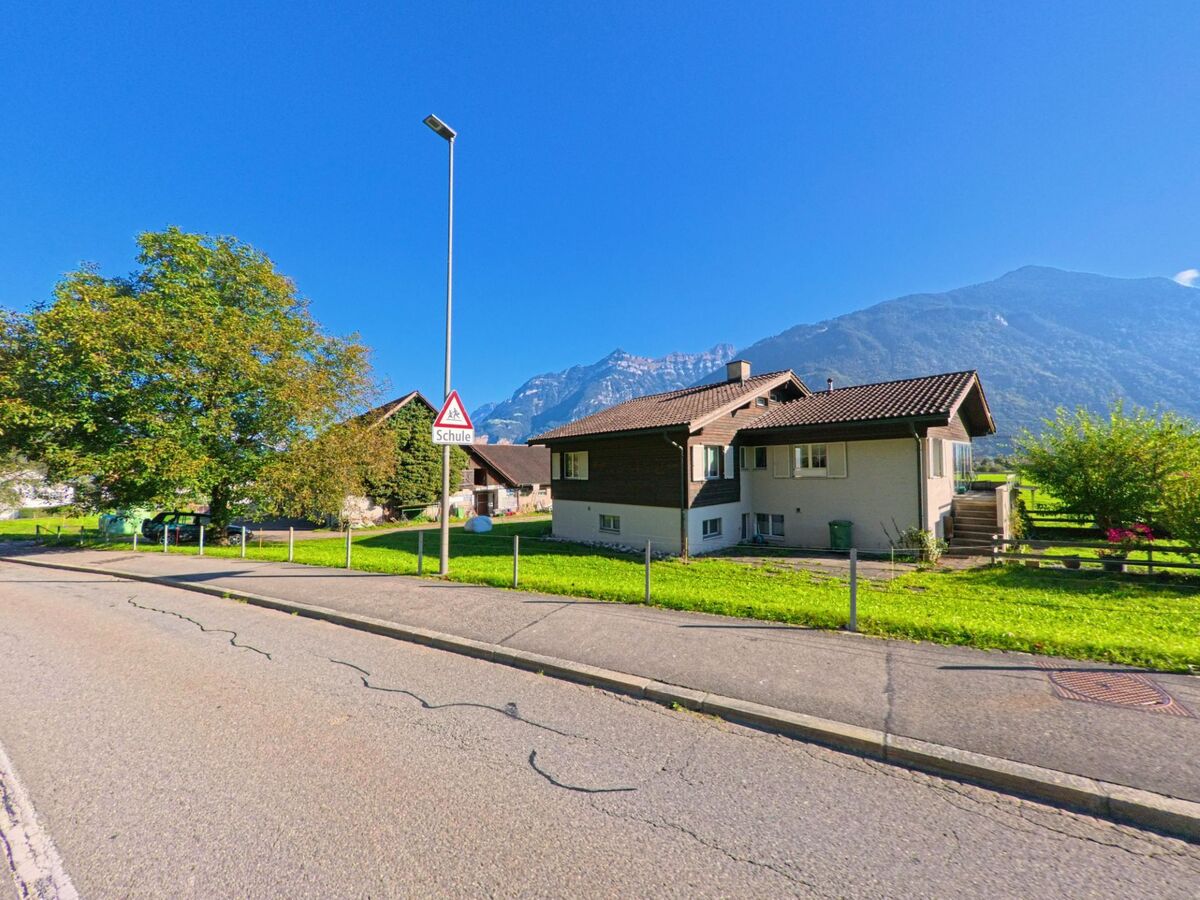 Bei einer Wohnstrasse mit Schulsignal stehen Einfamilienhäuser und Grünflächen in Seedorf (6462, Kanton Uri) vor einer weitläufigen Berglandschaft.