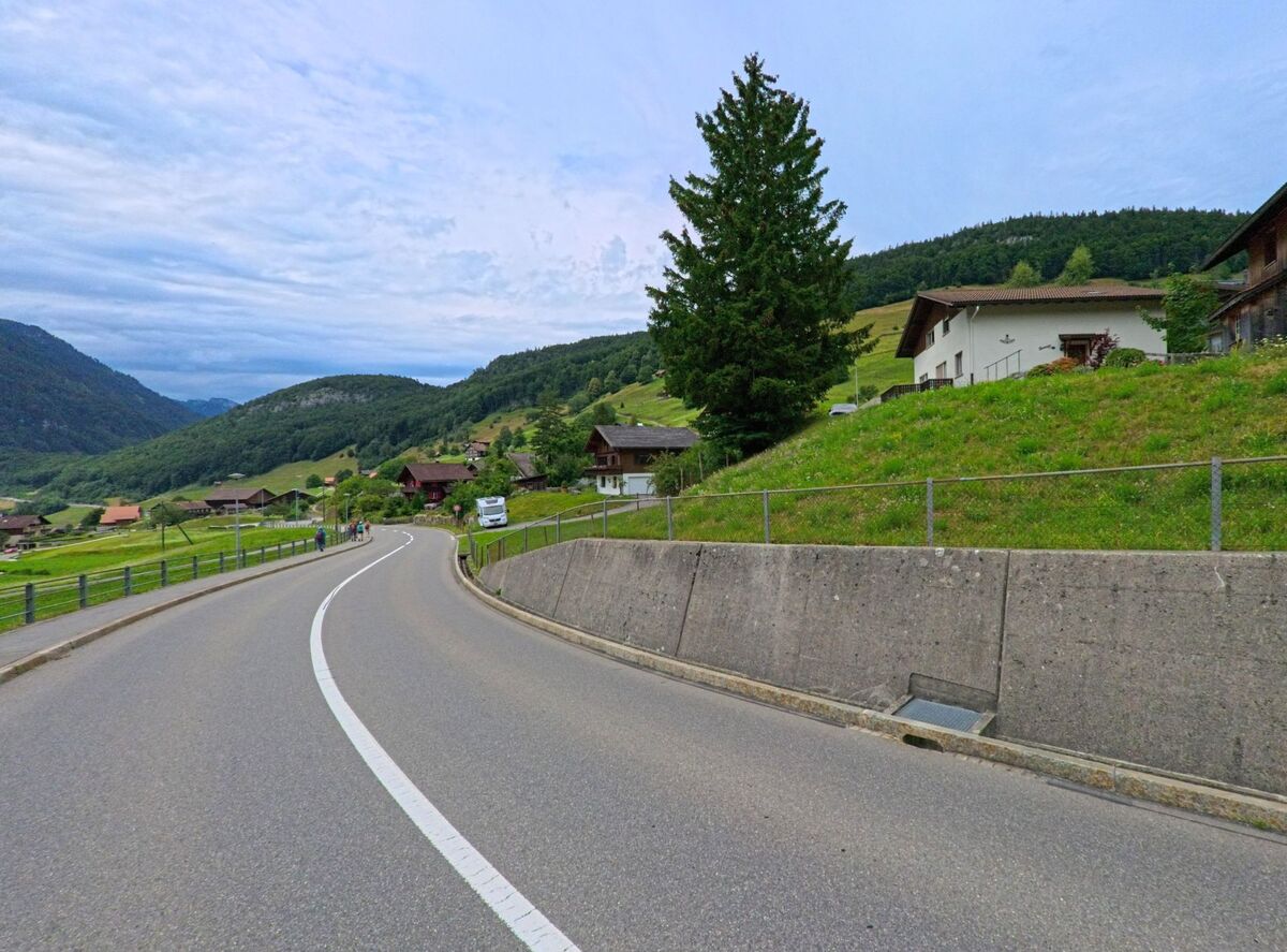 Eine geschwungene Strasse fuehrt in Seelisberg (6377, Kanton Uri) durch eine huegelige Landschaft mit verstreuten Wohnhaeusern und dichten Waldabschnitten.