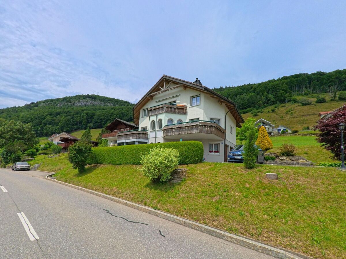 In Seelisberg (6377, Kanton Uri) steht ein gepflegtes Wohnhaus mit verziertem Balkon an einem Hang, umgeben von weiteren Haeusern und einem bewaldeten Bergruecken.