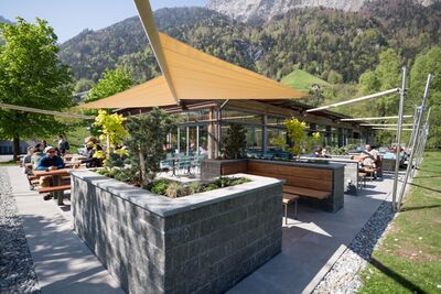 Gaeste sitzen in Seedorf UR (6462, Kanton Uri) auf der Terrasse eines modernen Restaurants unter grossen Sonnensegeln mit Blick auf die umliegenden Berge.