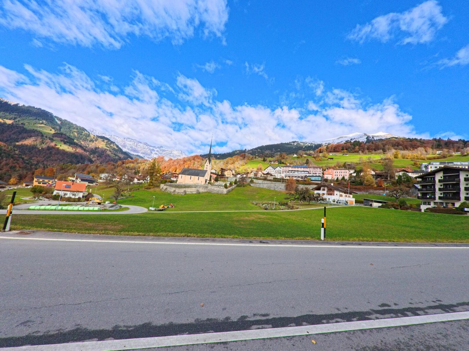 Über eine freie Wiesenfläche hinweg zeigt sich das Dorfzentrum von Seewis im Prättigau (7212, Kanton Graubünden) mit Kirche, Wohnhäusern und herbstlich gefärbten Hängen vor alpiner Kulisse.