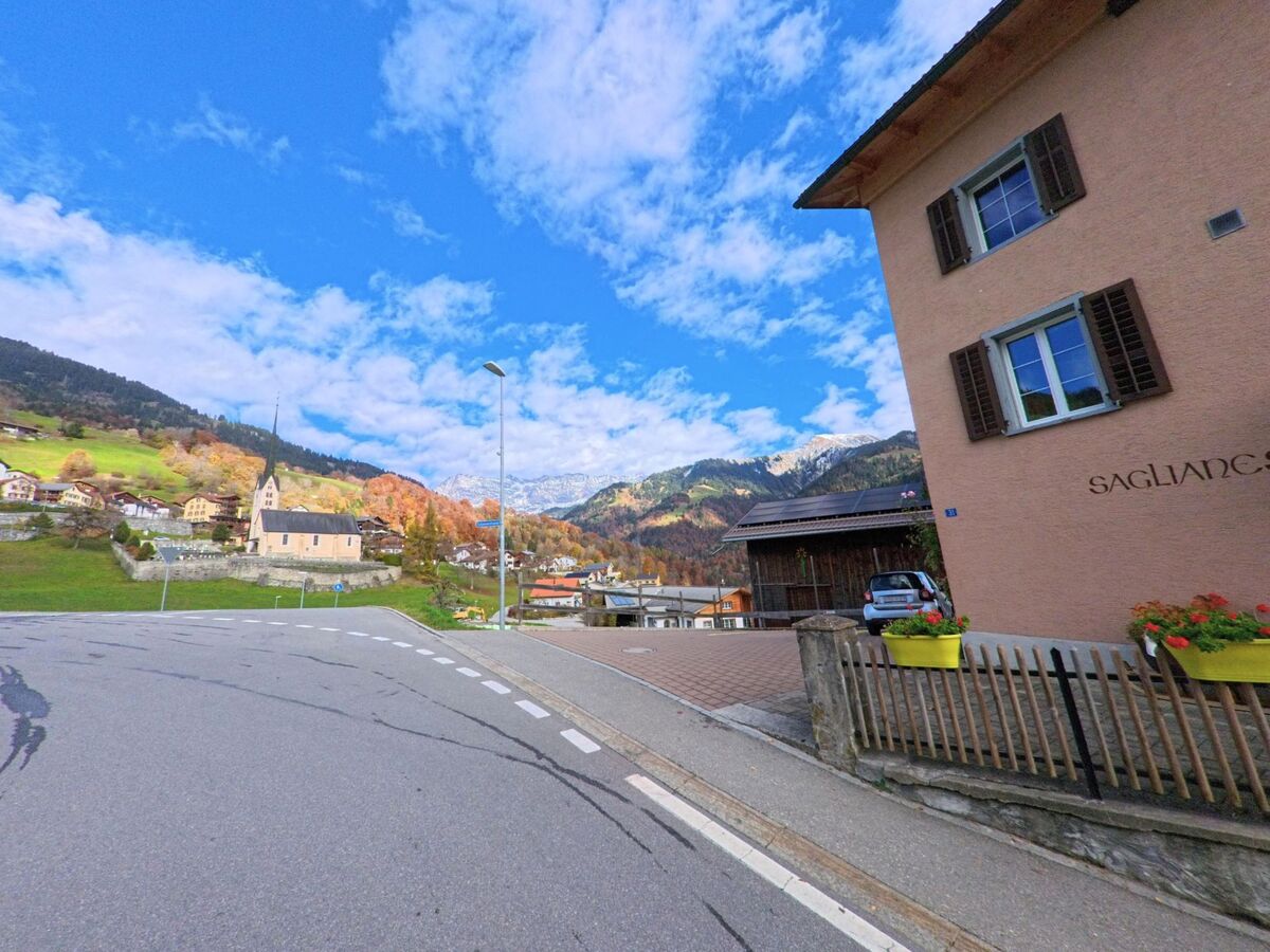 An einer sanft geschwungenen Strasse öffnet sich der Blick auf das Dorf Seewis im Prättigau (7212, Kanton Graubünden) mit Kirche, Wohnhäusern und herbstlich gefärbten Hängen vor alpiner Kulisse.