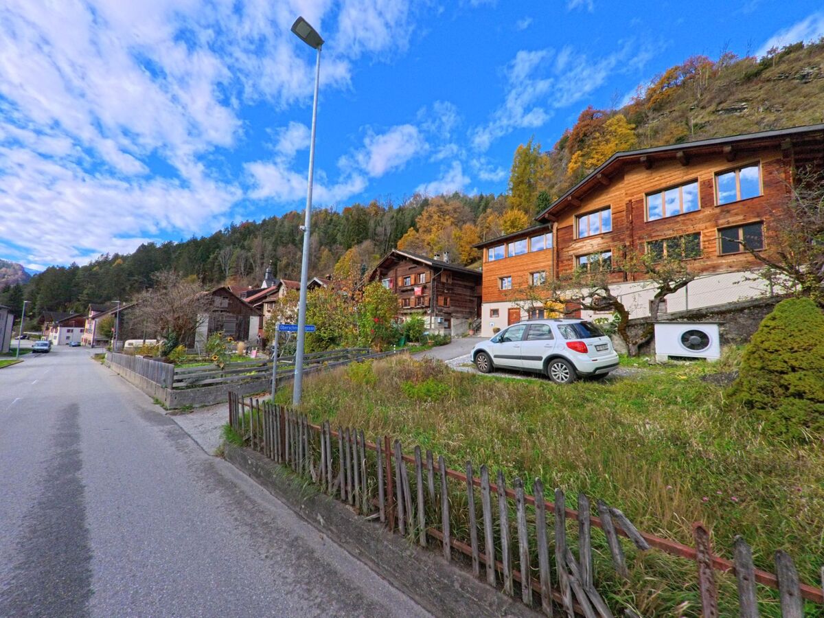 Entlang einer ruhigen Dorfstrasse stehen Holzhäuser und Gärten in Seewis im Prättigau (7212, Kanton Graubünden), eingebettet in einen herbstlich gefärbten Hang mit lockerem Baumbestand.