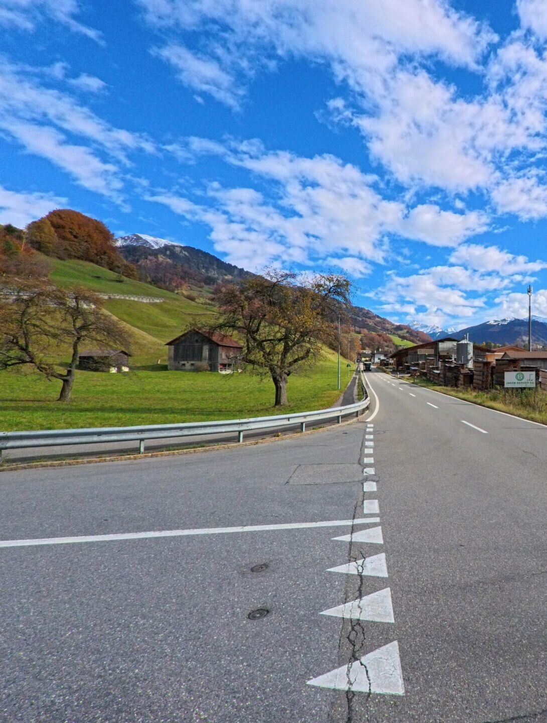 An einer markierten Kreuzung verläuft eine Strasse durch die offene Landschaft von Seewis im Prättigau (7212, Kanton Graubünden), flankiert von Wiesen, einzelnen Ställen und sanften Berghängen.