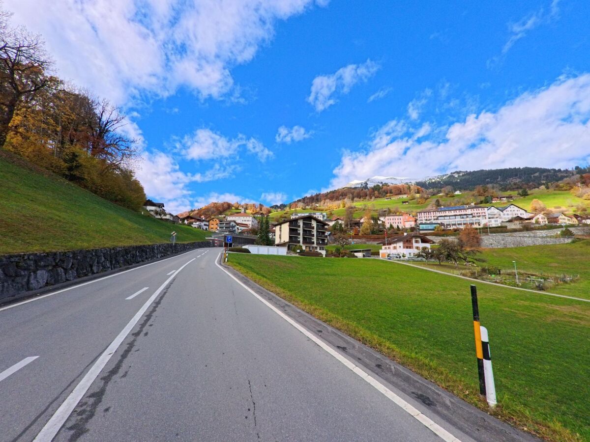 Entlang einer breiten Talstrasse führt der Blick auf das Siedlungsgebiet von Seewis im Prättigau (7212, Kanton Graubünden), eingerahmt von grünen Wiesen, Hanglagen und herbstlich gefärbten Bäumen.