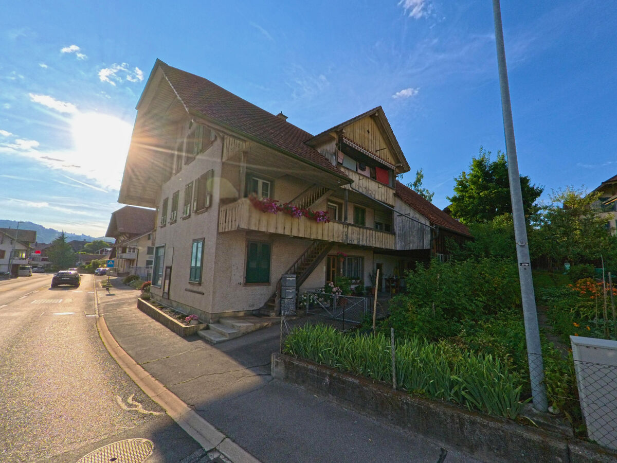 In Seftigen (3662, Kanton Bern) wirft die tiefstehende Sonne warmes Licht auf ein älteres Wohnhaus mit Holzveranda und kleinem Vorgarten an der Hauptstrasse.