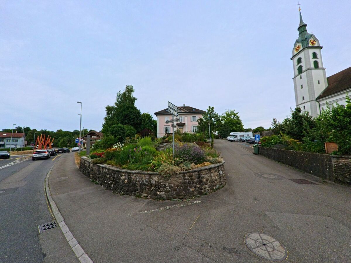 In Sempach (6204, Kanton Luzern) führt ein schmaler Weg an einem blühenden Gartenhang vorbei zur Kirche mit markantem Turm und Ziffernblättern.