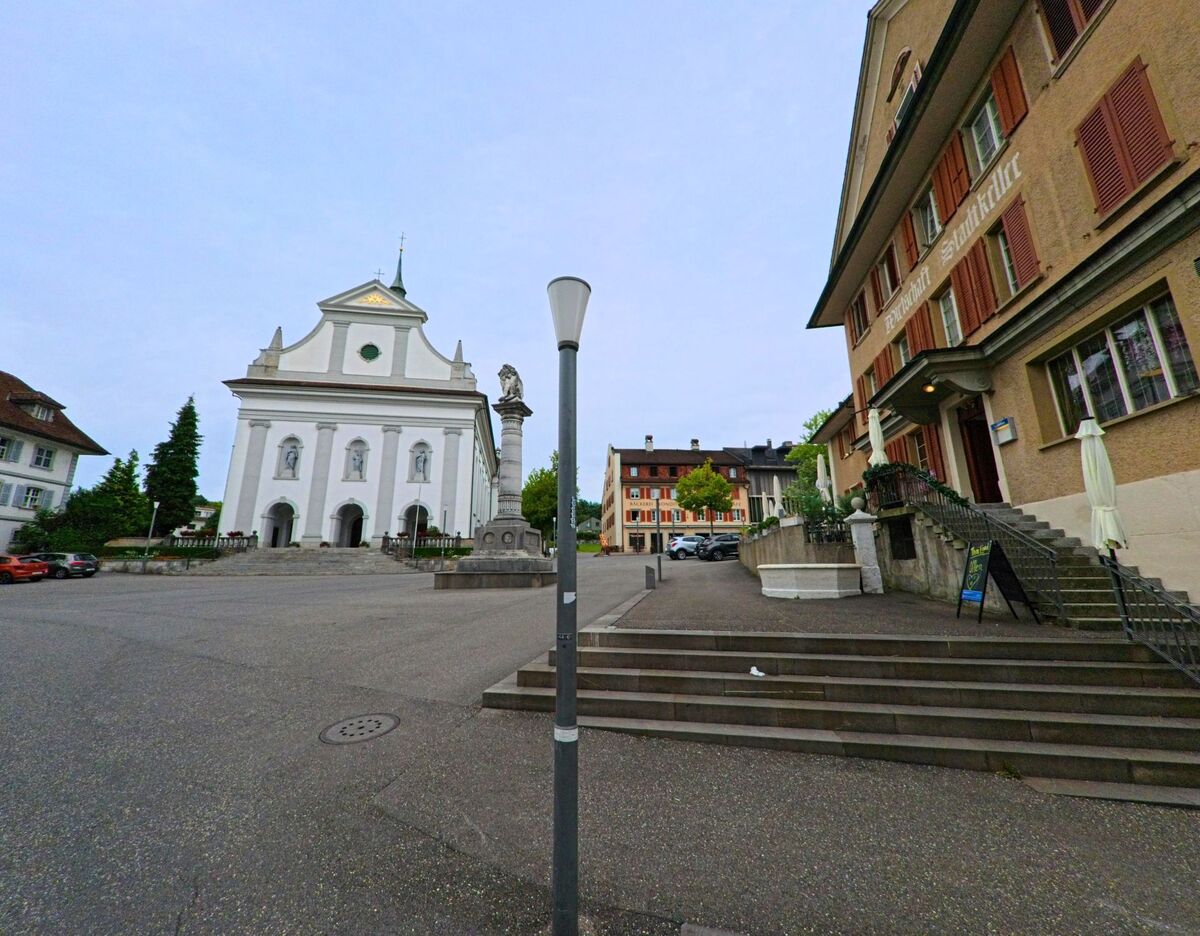 In Sempach (6204, Kanton Luzern) stehen sich die weisse Stadtkirche und das Gebäude der Wirtschaft Stadtkeller an einem weiten Platz gegenüber, in dessen Mitte eine steinerne Säule aufragt.