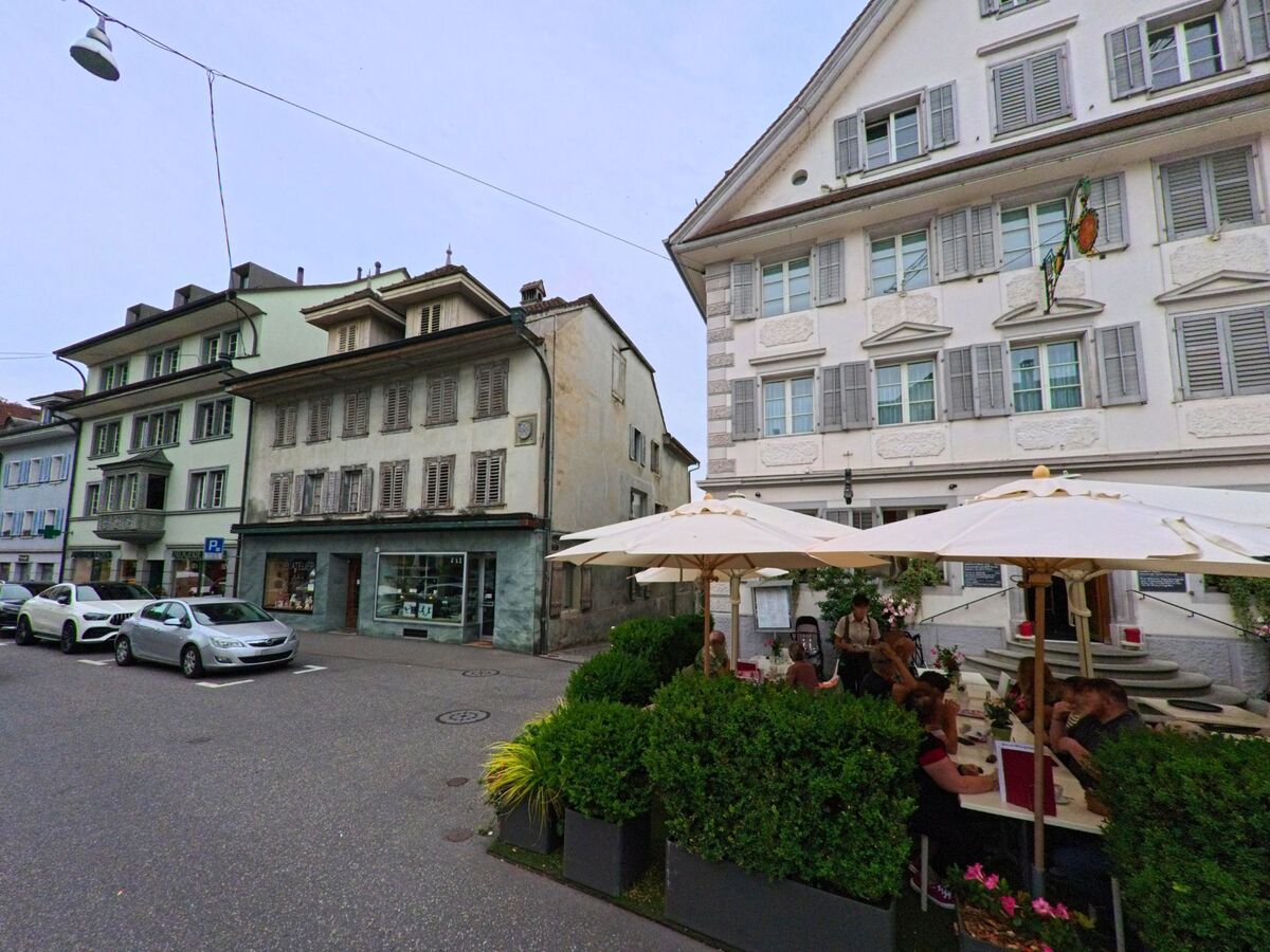 Im Zentrum von Sempach (6204, Kanton Luzern) sitzen Menschen unter weissen Sonnenschirmen vor einem traditionellen Gasthaus mit grauen Fensterläden.