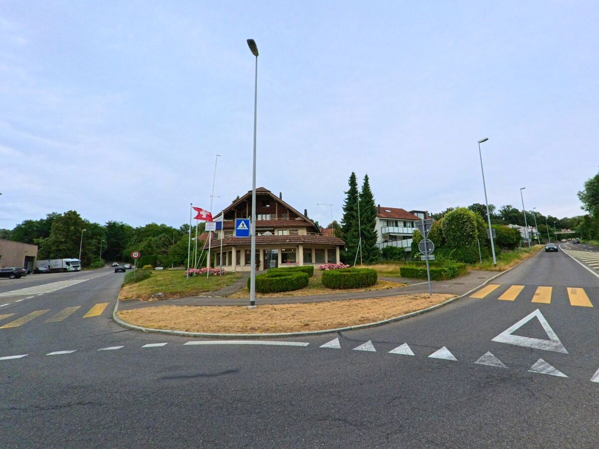Ein traditionelles Holzhaus mit Schweizer Fahne steht an einer Verkehrsinsel in Sempach (6204, Kanton Luzern), umgeben von Zebrastreifen und gepflegten Strassen.