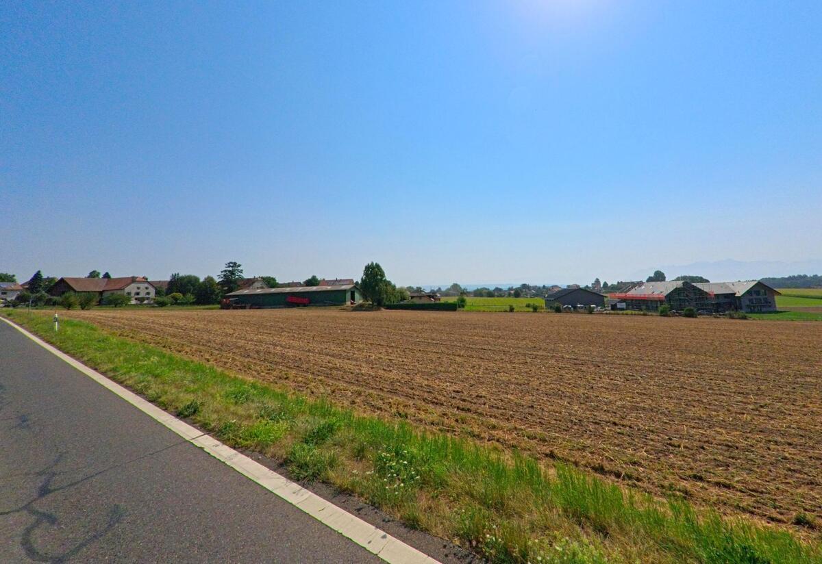 Von der Strasse aus blickt man in Senarclens (1308, Kanton Waadt) auf weite Ackerflaechen und mehrere landwirtschaftliche Gebaeude am Dorfrand.