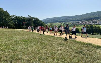 Auf einem breiten Kiesweg wandert in Muttenz (4132, Kanton Baselland) eine Gruppe von Seniorinnen und Senioren gemeinsam durch eine offene Landschaft mit Blick auf das Tal.