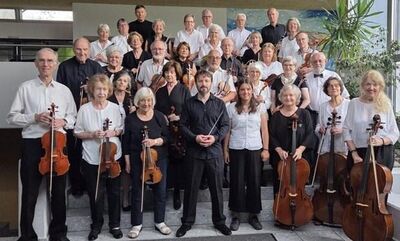 Das Seniorenorchester der Region Baden aus Frick (5070, Kanton Aargau) steht gemeinsam mit Instrumenten für ein Gruppenfoto in einem hellen Saal.