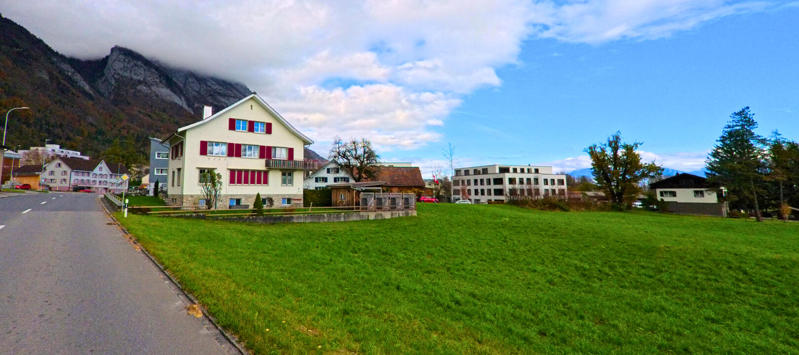 Am Rand einer Wohnsiedlung in Sennwald (9466, Kanton St. Gallen) liegen Einfamilienhaeuser und ein modernes Gebaeude neben einer weitlaeufigen Gruenflaeche vor Bergkulisse.