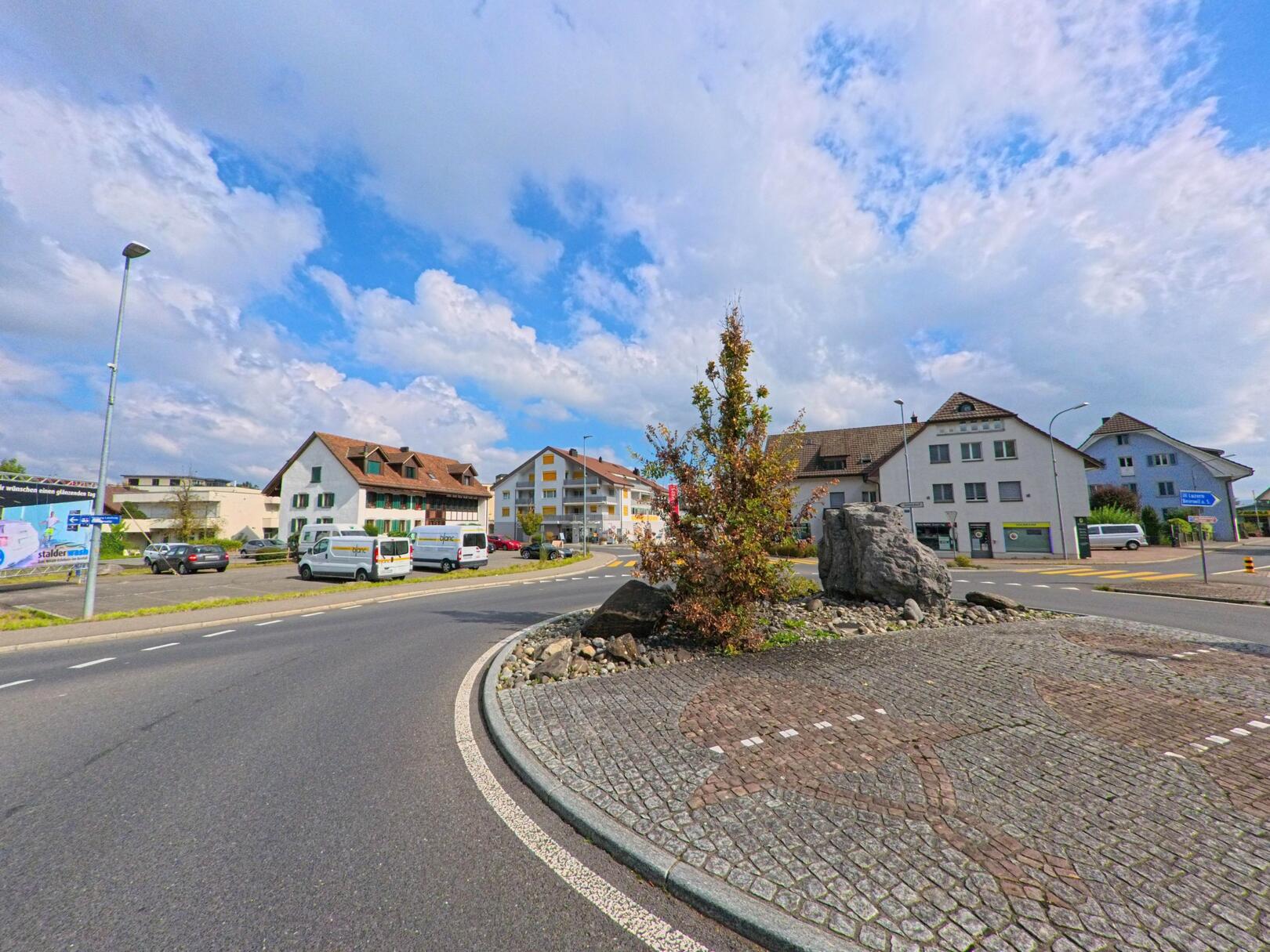 Am Kreisverkehr in Seon (5703, Kanton Aargau) treffen moderne und traditionelle Wohnhäuser unter einem teilweise bewölkten Himmel aufeinander.