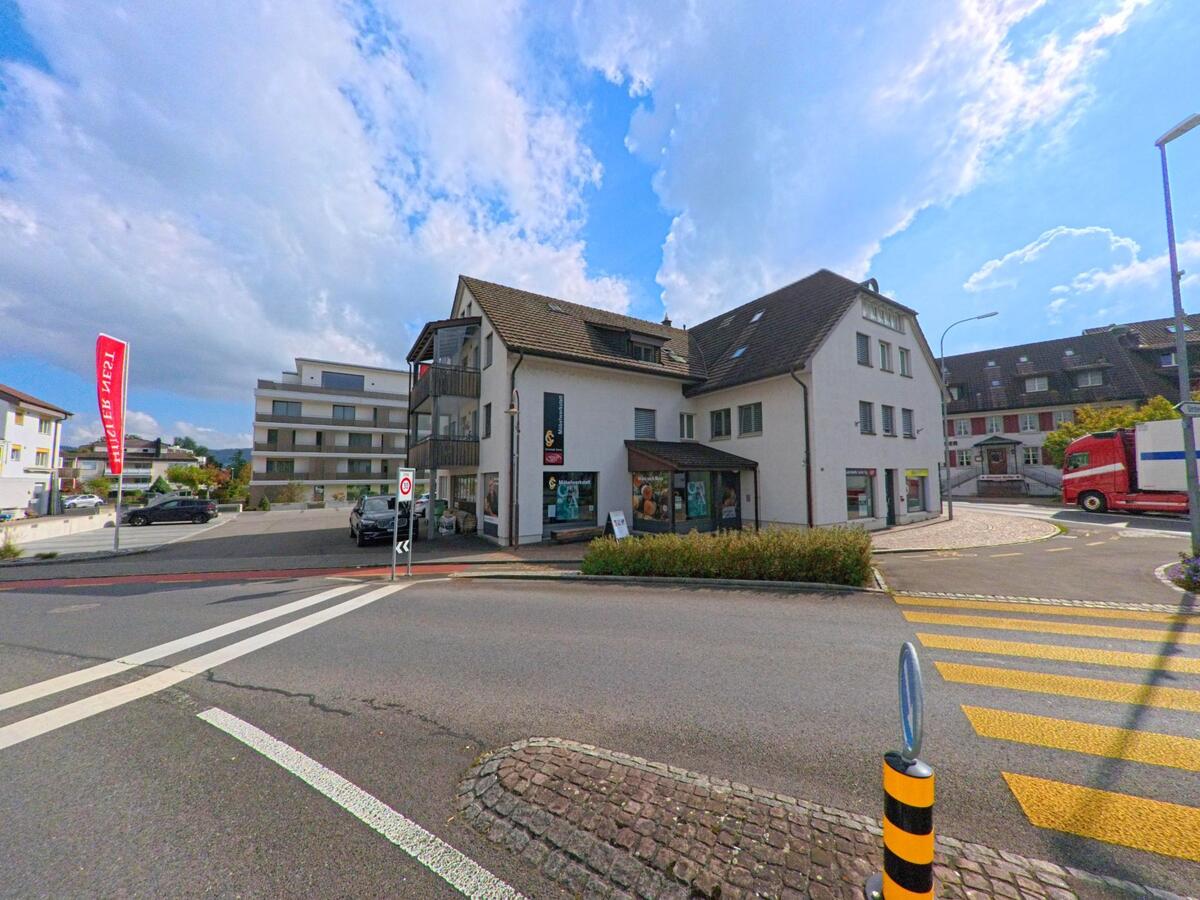 An einer belebten Kreuzung in Seon (5703, Kanton Aargau) stehen moderne Wohn- und Geschaeftshaeuser mit Parkplaetzen und Strassenmarkierungen unter einem hellen Himmel.