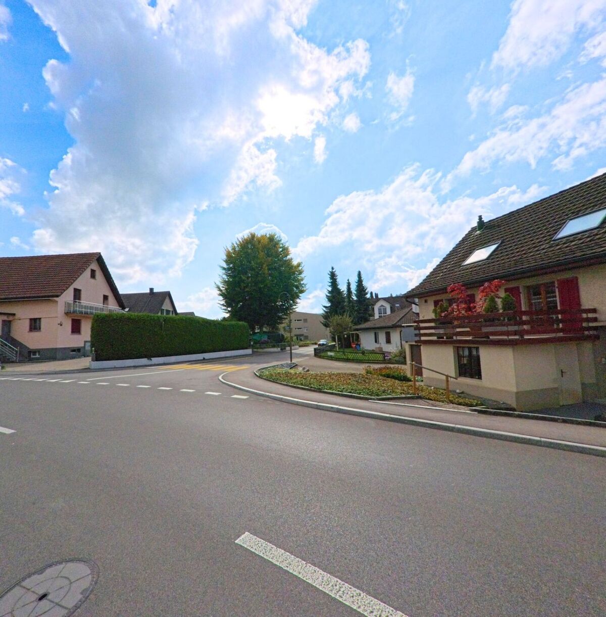 Eine ruhige Strasseneinmuendung in Seon (5703, Kanton Aargau) zeigt Einfamilienhaeuser mit gepflegten Gaerten und einer kleinen Verkehrsinsel unter leicht bewolktem Himmel.
