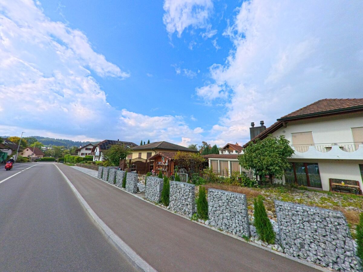 Entlang einer Wohnstrasse in Seon (5703, Kanton Aargau) stehen Einfamilienhaeuser mit gepflegten Gaerten und Steinmauern unter blauem Himmel.