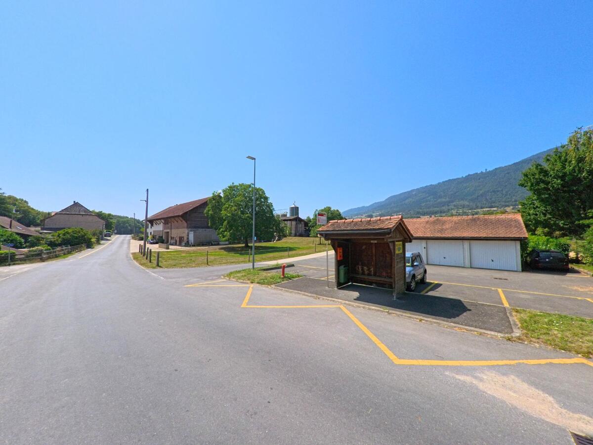 Eine kleine Bushaltestelle mit Holzunterstand steht an der Hauptstrasse in Sergey (1355, Kanton Waadt) neben Bauernhaeusern.