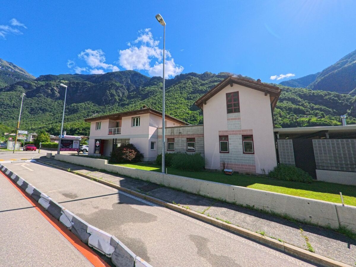 Entlang einer Strasse in Serravalle (6713, Kanton Tessin) stehen moderne Wohnhaeuser vor der Kulisse bewaldeter Berghaenge.