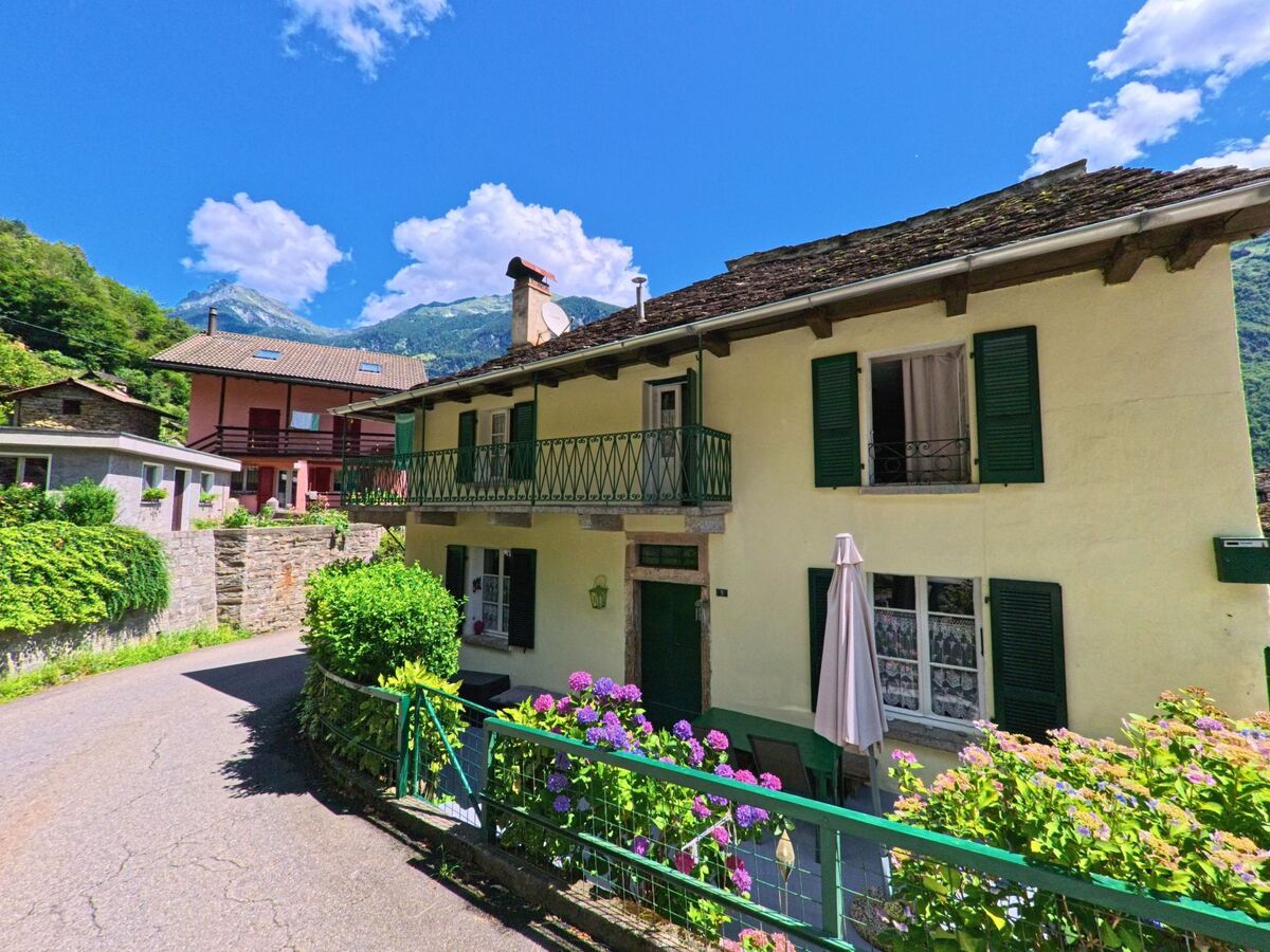 Ein gelbes Wohnhaus mit gruenen Fensterlaeden und blumengeschmuecktem Vorgarten steht in Serravalle (6714, Kanton Tessin) an einer schmalen Dorfstrasse.