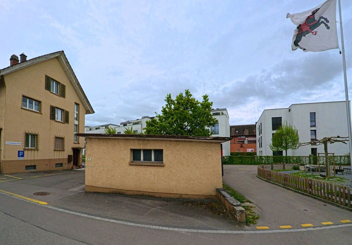 Zwischen älteren und neueren Wohngebäuden in Seuzach (8472, Kanton Zürich) steht ein flacher Anbau vor einem kleinen Platz mit Fahne und Sitzbänken.