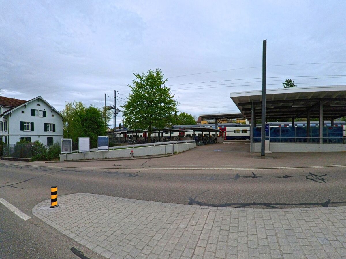 Ein Zug steht am Perron des Bahnhofs in Seuzach (8472, Kanton Zürich), daneben sind ein gedeckter Fahrradabstellplatz und ein Wohnhaus zu sehen.