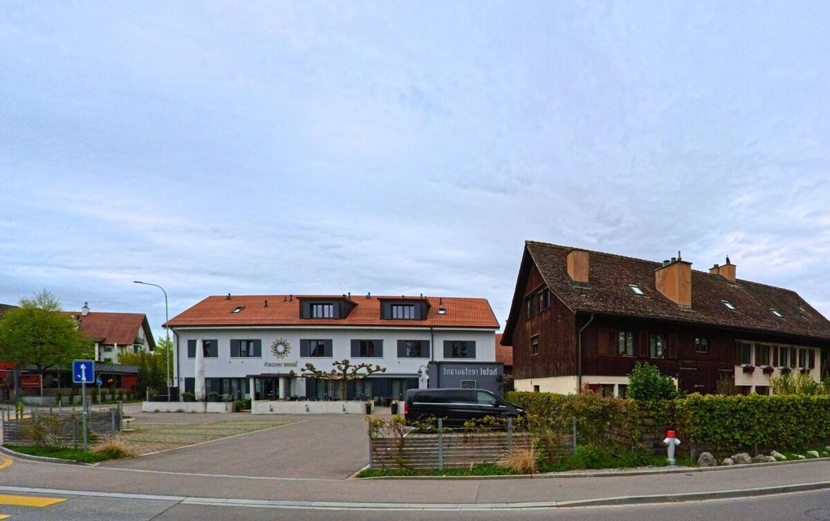 In Seuzach (8472, Kanton Zürich) stehen ein modernes Hotel-Restaurant mit heller Fassade und ein traditionelles Holzhaus Seite an Seite entlang einer Hauptstrasse.