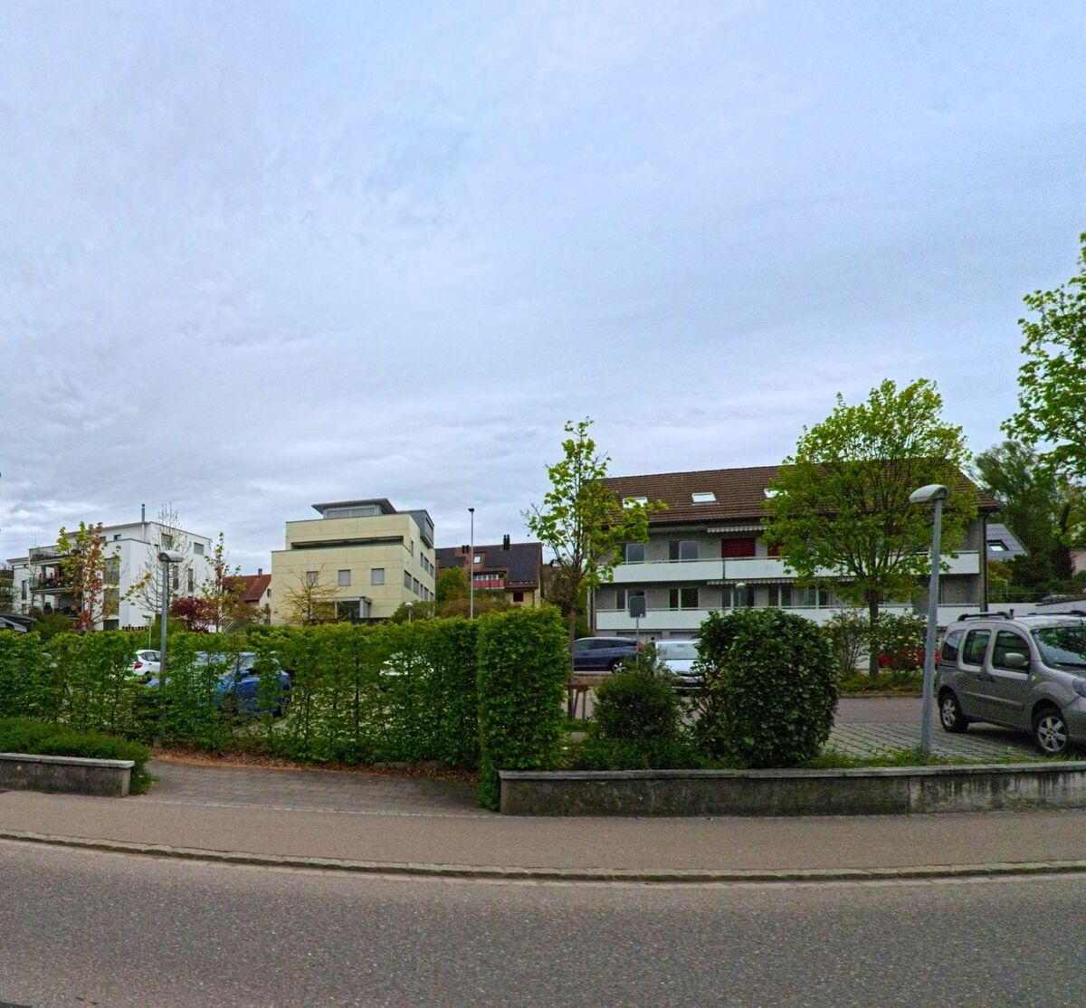 In Seuzach (8472 im Kanton Zürich) stehen moderne und traditionelle Mehrfamilienhäuser hinter einer gepflegten Hecke mit angrenzendem Parkplatz.