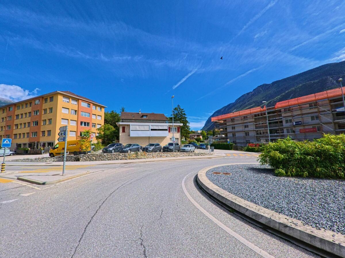 Eine kurvige Strasse fuehrt an Wohnhaeusern in Sierre (3960, Kanton Wallis) vorbei, im Hintergrund sind eingeruestete Gebaeude und die Berge sichtbar.