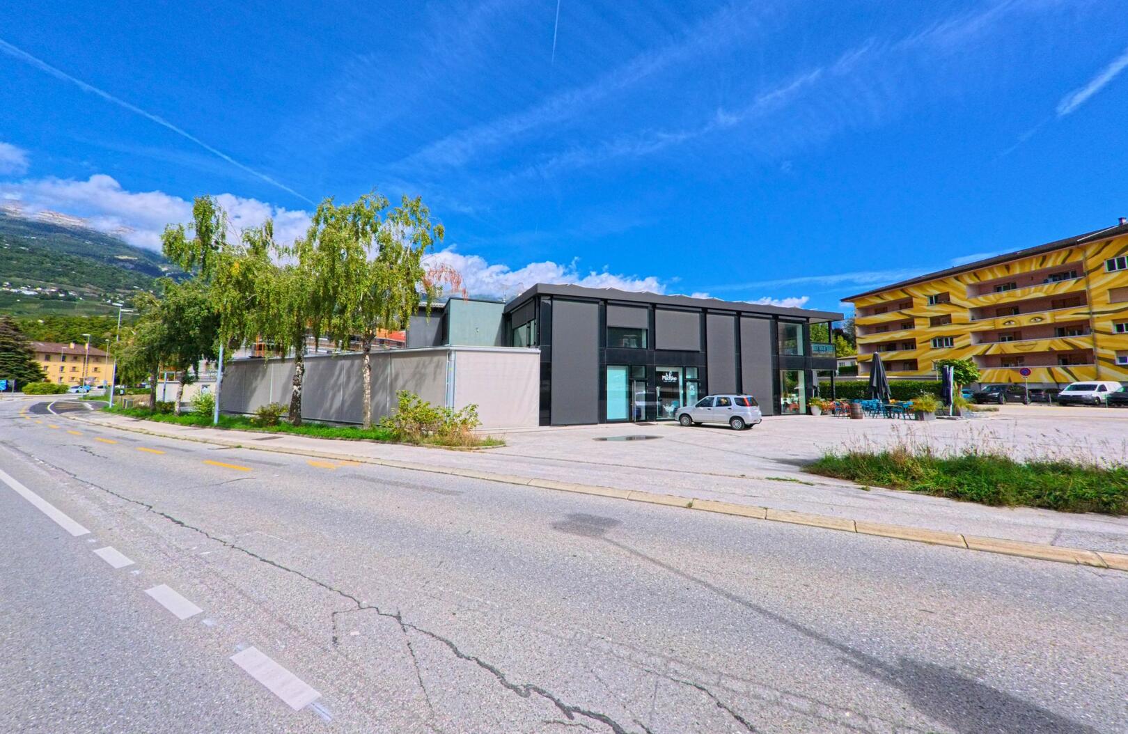 Ein modernes schwarzes Gebaeude mit grossen Glasfronten steht in Sierre (3960, Kanton Wallis) an einer breiten Strasse, daneben befinden sich Wohnhaeuser.